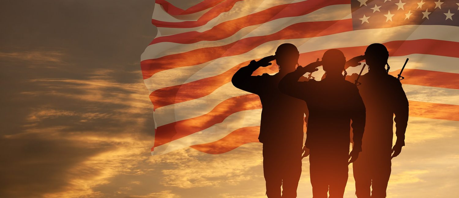 Three soldiers saluting in silhouette in front of an American flag at sunset
